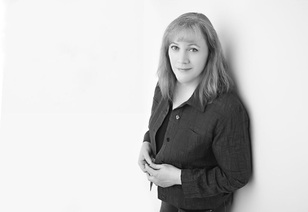 Black and white photo of a woman with light hair, wearing a dark patterned jacket over a top. Perfect for author headshots, she stands against a plain white wall, looking at the camera with a slight smile, hands holding her open jacket. - Jodi O Photography