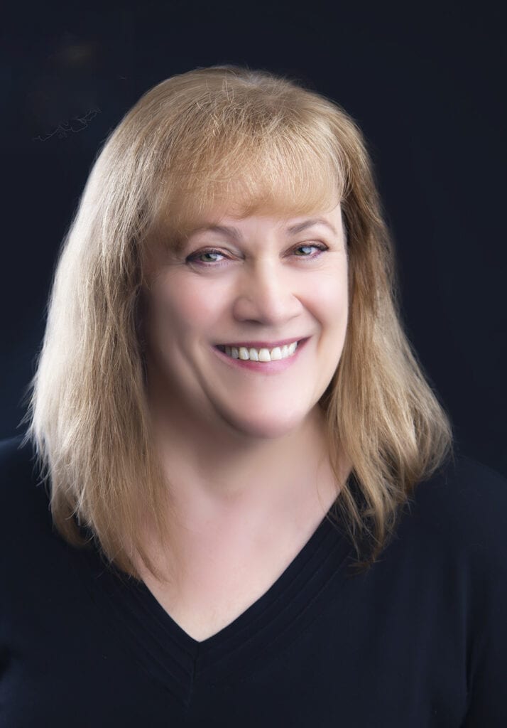 A woman with straight, shoulder-length blonde hair smiles at the camera. She is wearing a black top and is posed against a dark background, capturing the professional style often delivered by Calgary photographers for author headshots. - Jodi O Photography
