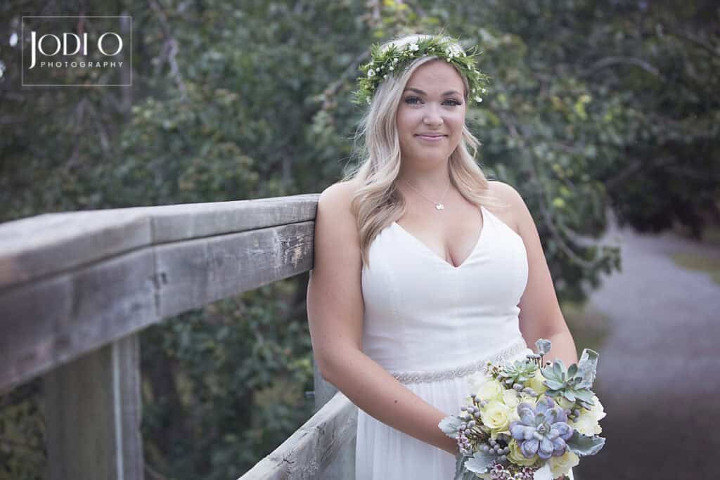 A bride in a white dress and floral crown stands smiling by a wooden railing outdoors, holding a bouquet of white and green flowers. Trees and a path are visible behind her, capturing the charm of Calgary couples with timeless wedding photography. - Jodi O Photography