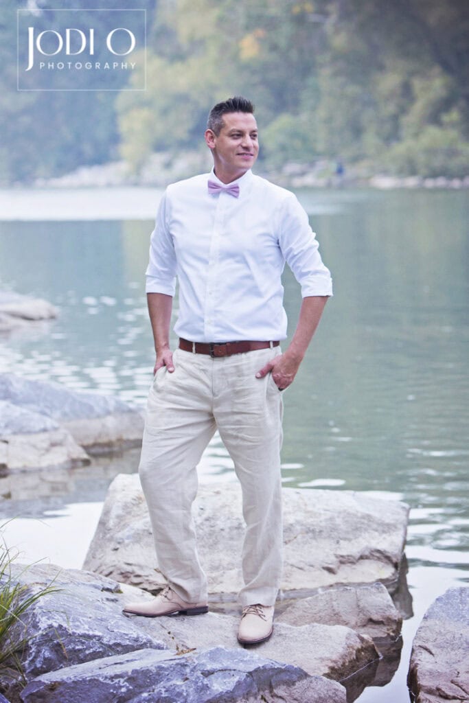 A man in light-colored pants, a white shirt, and a pink bow tie stands on large rocks by a calm river near Calgary, with trees in the background. The logo “JODI O PHOTOGRAPHY” is in the top left corner. - Jodi O Photography