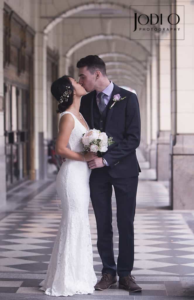 A bride in a white lace dress and groom in a dark suit kiss under an arched walkway. Captured by Calgary wedding photography, the bride holds a bouquet as they stand on a black-and-white geometric patterned floor. - Jodi O Photography