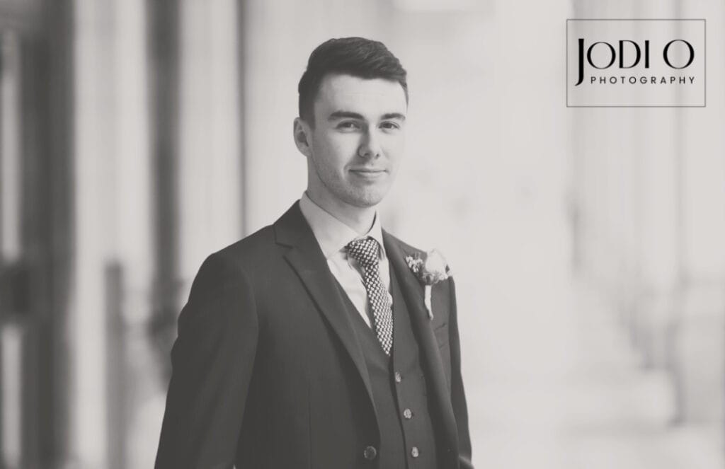 A young man in a suit and tie stands indoors, facing the camera and smiling slightly. The background is blurred, with the "Jodi O Photography" logo—specializing in Calgary wedding photography—in the top right corner. The image is in black and white. - Jodi O Photography