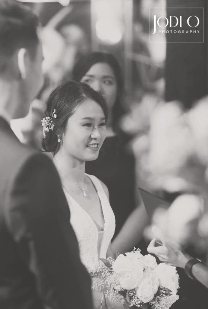A bride in a white dress holding a bouquet smiles at her groom during a Calgary wedding photography ceremony, with guests in the background. The image is in black and white and features the Jodi O Photography logo. - Jodi O Photography