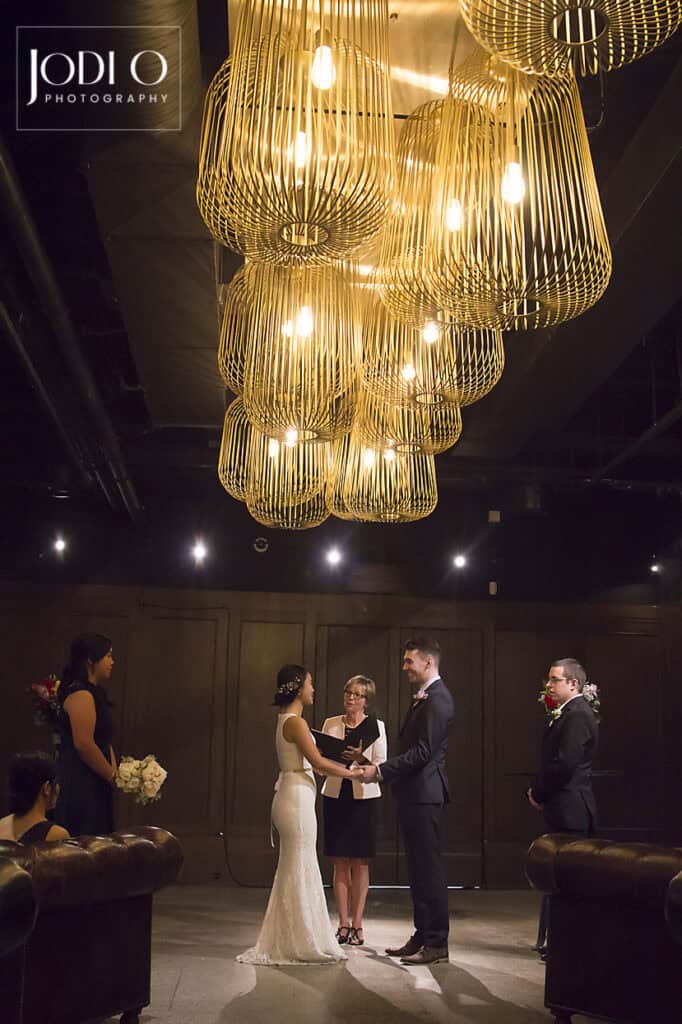 A bride and groom hold hands during their wedding ceremony under golden chandeliers, with a celebrant and two attendants nearby in a dimly lit, elegant room—perfect for timeless Calgary wedding photography. - Jodi O Photography
