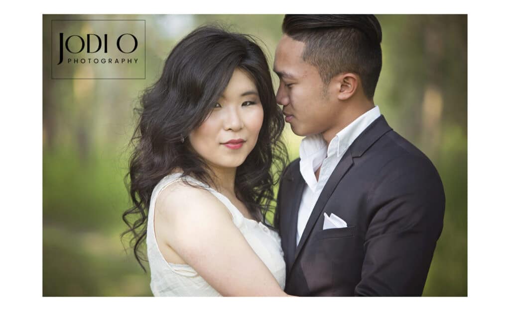 A couple stands close together outdoors in Calgary; the woman looks at the camera while the man faces her, smiling. Both are dressed formally, highlighting timeless wedding photography. The background is blurred greenery. "Jodi O Photography" logo is in the top left corner. - Jodi O Photography