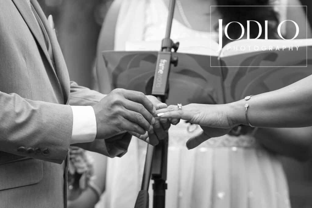 A groom places a ring on the bride’s finger during a Calgary wedding ceremony, with a microphone and officiant in the background. The black-and-white image by JODIO PHOTOGRAPHY beautifully captures this moment of wedding photography. - Jodi O Photography