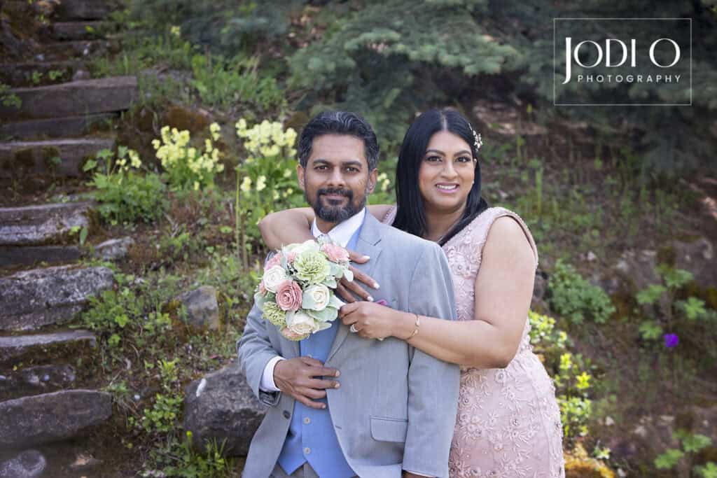 A couple poses outdoors in Calgary; the woman in a light pink dress hugs the man in a gray suit while holding a pastel bouquet. Near stone steps and greenery, they showcase elegant wedding photography by JODI O PHOTOGRAPHY. Hourly rate available. - Jodi O Photography