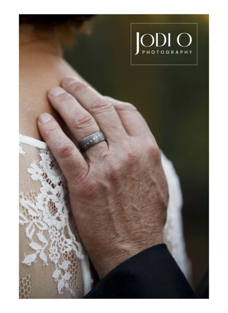 A close-up of a hand with a gray ring gently resting on a lace-covered shoulder, showcasing the artistry of Calgary wedding photography, with "Jodi O Photography" written in the top right corner. - Jodi O Photography