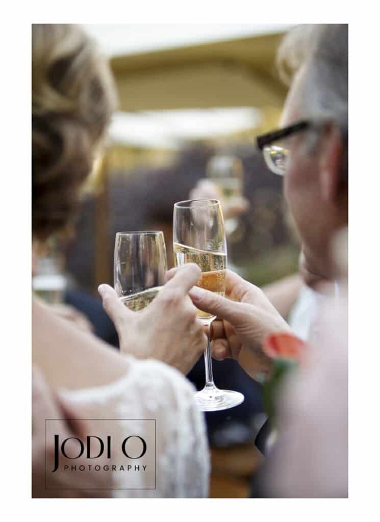 Two people clink champagne glasses in a celebratory toast, seen from behind at close range. Perfect for wedding photography in Calgary, the softly blurred background adds charm. Photo credited to Jodi O Photography. - Jodi O Photography