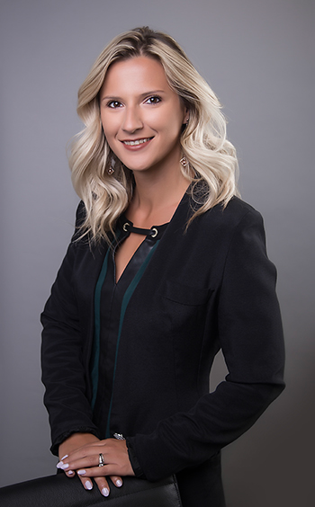 A woman with wavy blonde hair, wearing a black blazer over a dark top, smiles at the camera while standing with her hands resting on the back of a chair against a plain gray background—an ideal professional headshot for branding. - Jodi O Photography