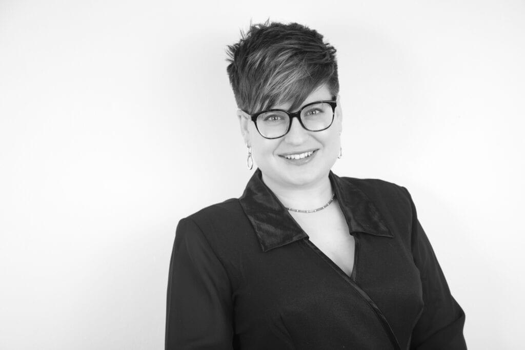 Black and white photo of a smiling person with short, dark hair and glasses, dressed in a dark blazer and light jewelry—exuding Portraits Success—standing against a plain white background. Perfect for Calgary Headshots or professional portraits. - Jodi O Photography