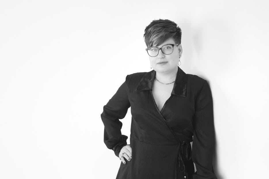 Black and white photo of a person with short, styled hair and glasses, wearing a dark wrap dress, stands against a plain white background with one hand on their hip—an ideal choice for Calgary Headshots and professional portraits. - Jodi O Photography