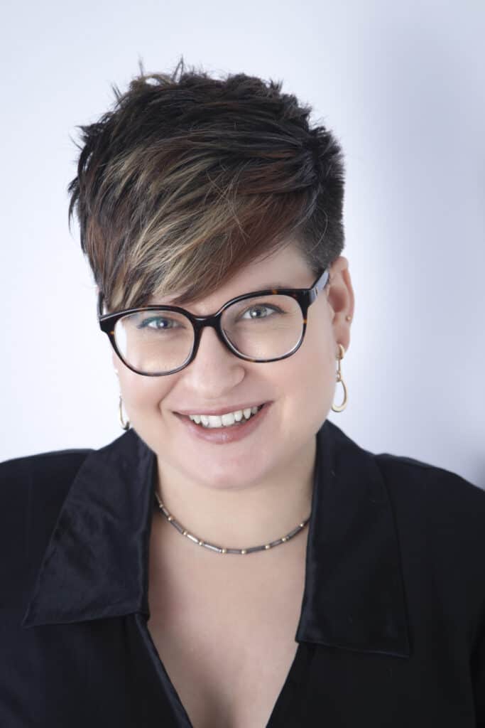 A smiling person with short, dark hair featuring light highlights, wearing black-framed glasses, gold hoop earrings, a beaded necklace, and a black collared top—capturing Portraits Success in front of a plain light background. - Jodi O Photography