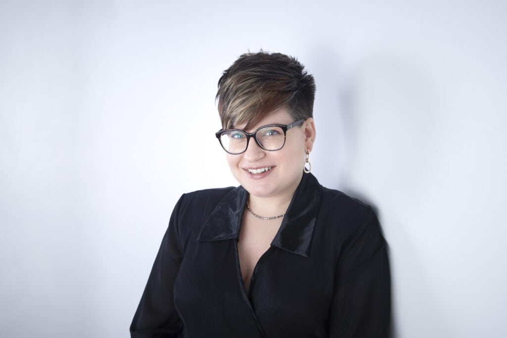 Person with short, layered hair and glasses, smiling and wearing a black shirt, stands against a plain white background—an ideal example of Calgary Headshots and portrait success. - Jodi O Photography