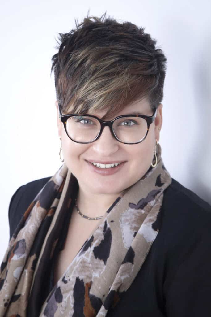 A person with short, layered brown hair and glasses smiles at the camera. Showcasing Calgary Headshots style, they wear hoop earrings, a patterned scarf, and a black top against a light background. - Jodi O Photography