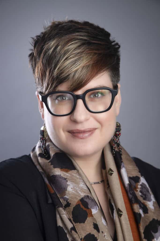 A person with short, brown and blonde hair, wearing black glasses, a patterned scarf, and beaded earrings, smiles slightly against a gray background—capturing the confidence sought in Calgary Headshots. - Jodi O Photography