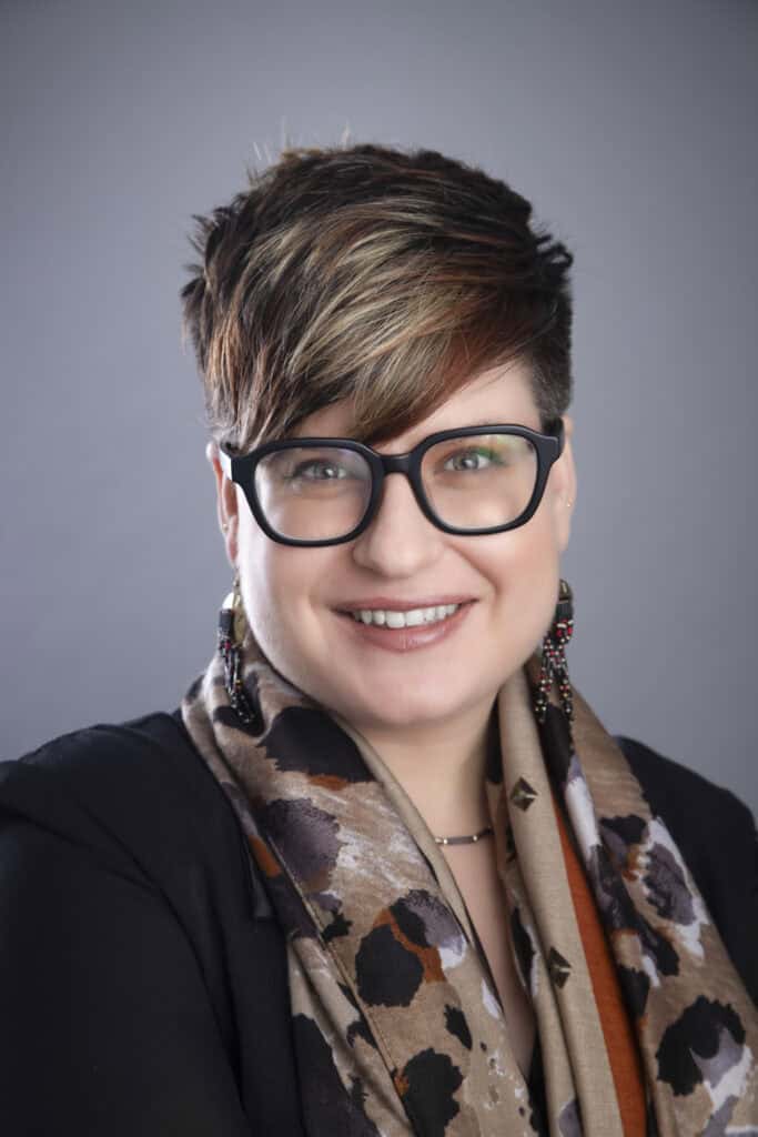 A person with short, layered brown hair, wearing large black glasses, a patterned scarf, and earrings, smiles in front of a gray background—an ideal example of Calgary Headshots and Professional Portraits for Portraits Success. - Jodi O Photography