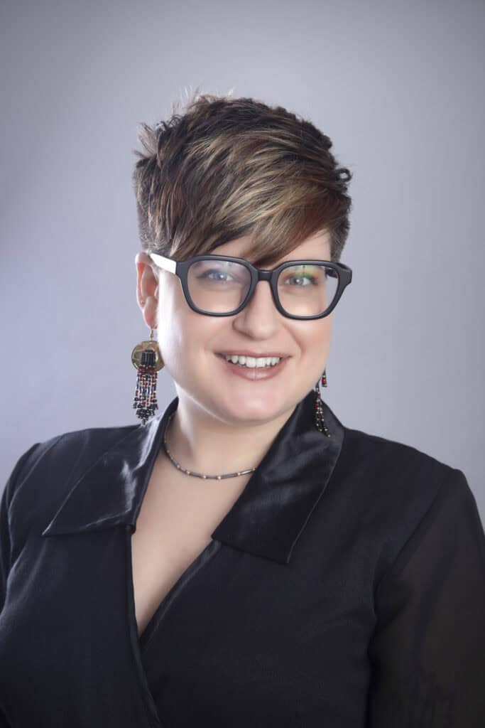 A person with short, layered hair and glasses smiles at the camera. Showcasing Calgary Headshots style, they wear large beaded earrings, a black collared top, and a thin necklace against a neutral gray background. - Jodi O Photography