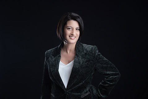 A woman with short dark hair, wearing a dark patterned blazer over a white blouse, stands against a black background, smiling with one hand on her hip—perfect for Calgary Headshots and professional branding. - Jodi O Photography