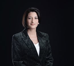 A woman with short dark hair wearing a patterned dark blazer over a white top poses confidently against a plain black background, capturing the essence of professional headshots ideal for modern branding. - Jodi O Photography