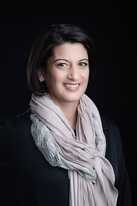 A woman with short dark hair smiles at the camera, wearing a black top and a textured scarf. Her polished look against a dark background exemplifies Calgary Headshots for professional branding. - Jodi O Photography