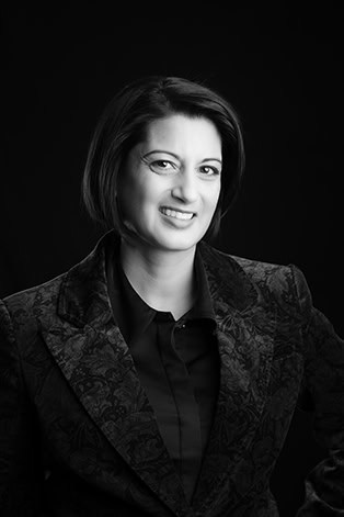 Black and white portrait of a woman with short dark hair, smiling in a patterned blazer over a dark shirt, posing confidently against a plain dark background—perfect for professional headshots or branding in Calgary. - Jodi O Photography