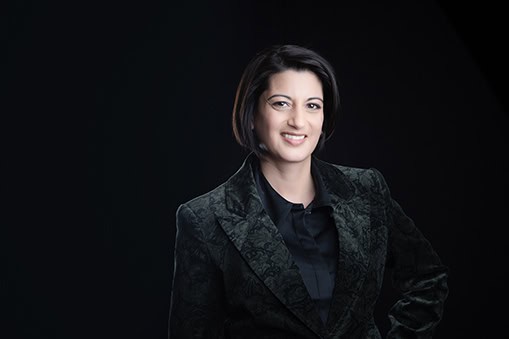 A woman with short dark hair, wearing a dark patterned blazer and a black shirt, smiles confidently against a plain dark background—ideal for professional headshots or impactful branding. - Jodi O Photography