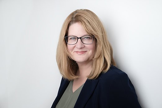 A woman with shoulder-length blonde hair and glasses smiles while wearing a navy blazer and green top, standing against a plain white background—ideal for professional business photos to enhance your brand. - Jodi O Photography