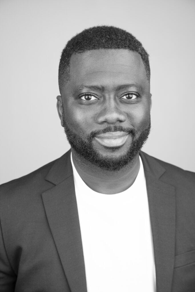 Black man with short hair and a beard wearing a dark blazer over a white shirt, smiling softly at the camera against a plain, light background—perfect for professional business photos to enhance your brand. - Jodi O Photography