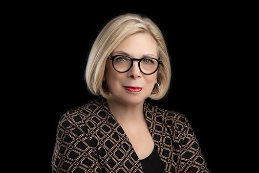 A woman with short blonde hair and red lipstick wears round glasses and a patterned black jacket, posing against a solid black background—perfect for professional business photos to enhance your brand. - Jodi O Photography
