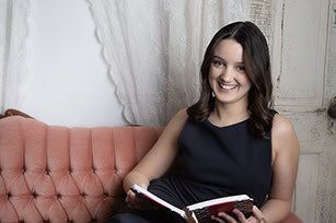 A woman with dark hair in a sleeveless black top sits on a pink tufted sofa, smiling while holding an open book. Behind her are white curtains and a rustic white door—perfect for professional business photos to enhance your brand. - Jodi O Photography