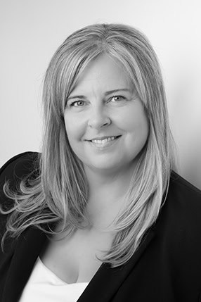 Black and white portrait of a smiling woman with long, straight, light-colored hair in a dark blazer over a light top, posed against a plain background—ideal for professional business photos and brand enhancement. - Jodi O Photography