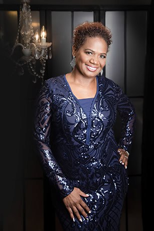 A woman smiles confidently, standing with one hand on her hip in a navy blue sequined dress. With short curly hair, she poses before a black paneled backdrop and lit chandelier—perfect for professional business photos to enhance your brand. - Jodi O Photography
