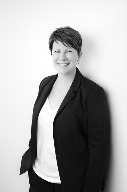 Black and white professional business photo of a person with short hair in a dark blazer over a light top, smiling and standing against a plain light background with one hand in their pocket. Perfect for brand enhancement. - Jodi O Photography