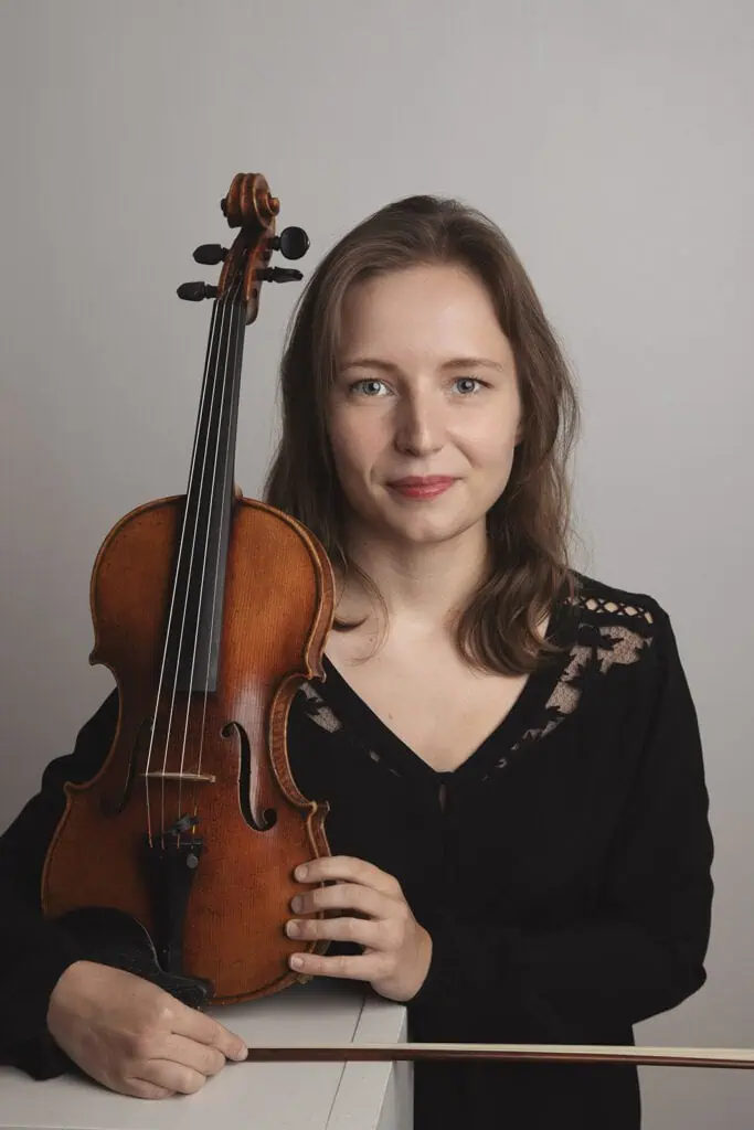 A woman with light brown hair, wearing a black top, smiles softly while holding a violin upright in front of her against a plain, light gray background—an image perfect for professional business photos to enhance your brand. - Jodi O Photography
