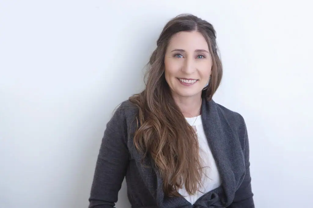 A woman with long brown hair, wearing a dark gray cardigan over a white top, smiles while standing against a plain, light-colored background—perfect for professional business photos to enhance your brand. - Jodi O Photography