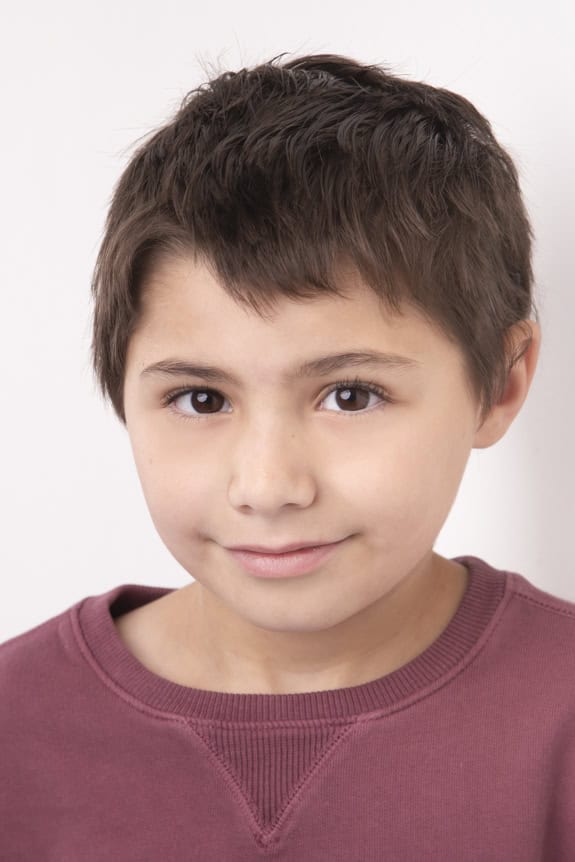 A young boy with short brown hair and brown eyes smiles softly, wearing a maroon crew-neck sweatshirt against a plain white background—an ideal example of Calgary kids’ headshots. - Jodi O Photography
