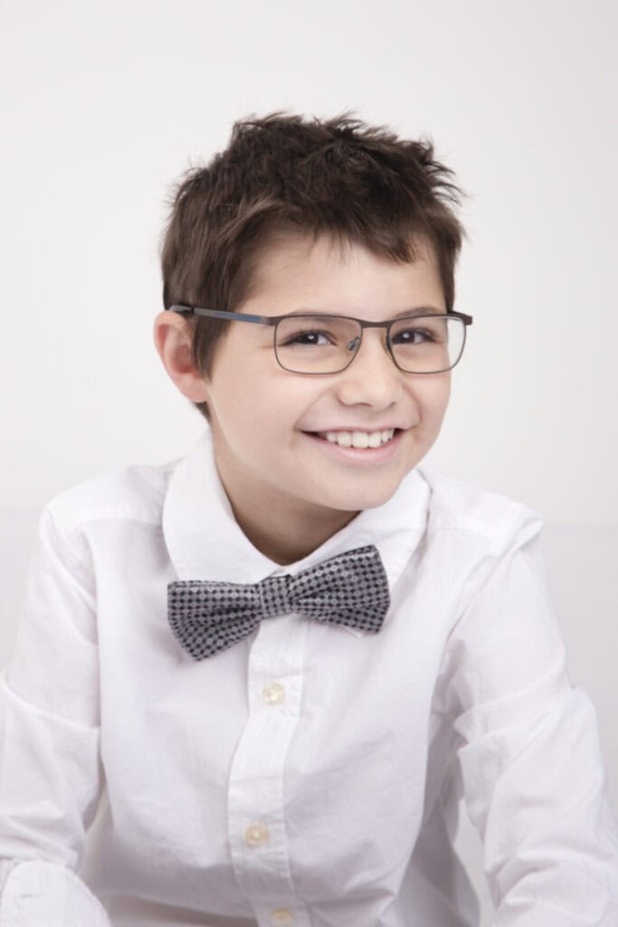 A smiling young boy with short brown hair, wearing glasses, a white shirt, and a grey bow tie, sits against a plain light background—perfect for amazing kids headshots in Calgary. - Jodi O Photography