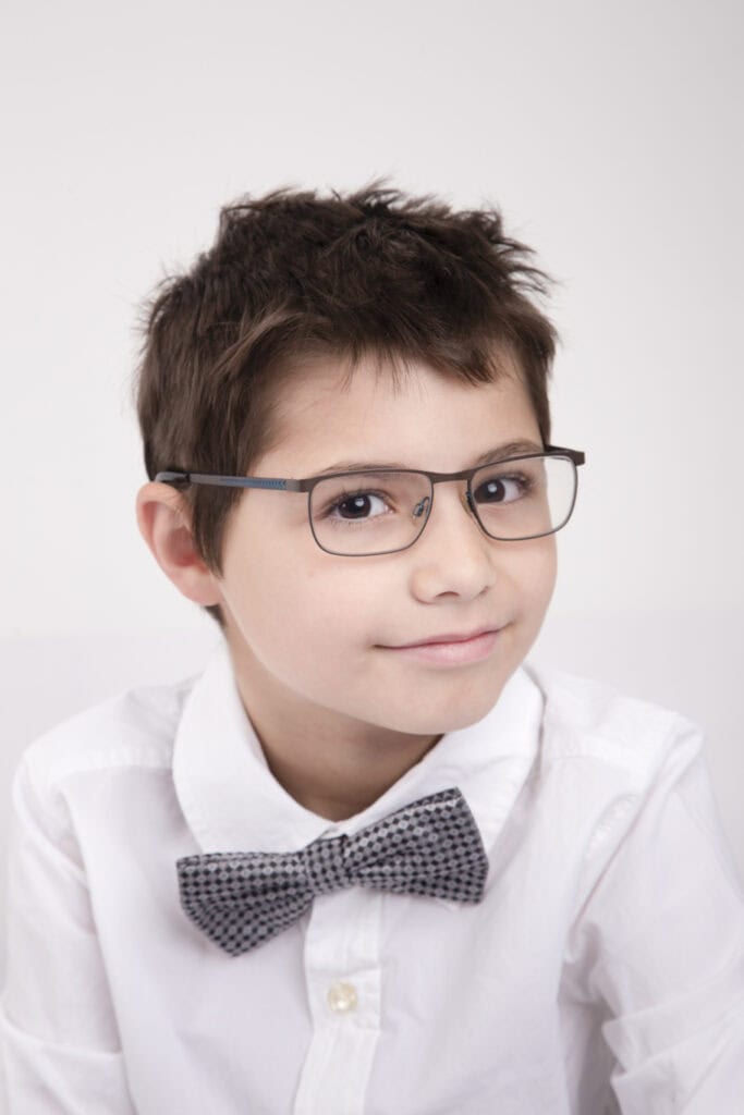 A young boy with short brown hair wearing glasses, a white button-up shirt, and a patterned bow tie, smiling slightly against a plain light background—perfect for kids headshots Calgary photographers love to capture. - Jodi O Photography