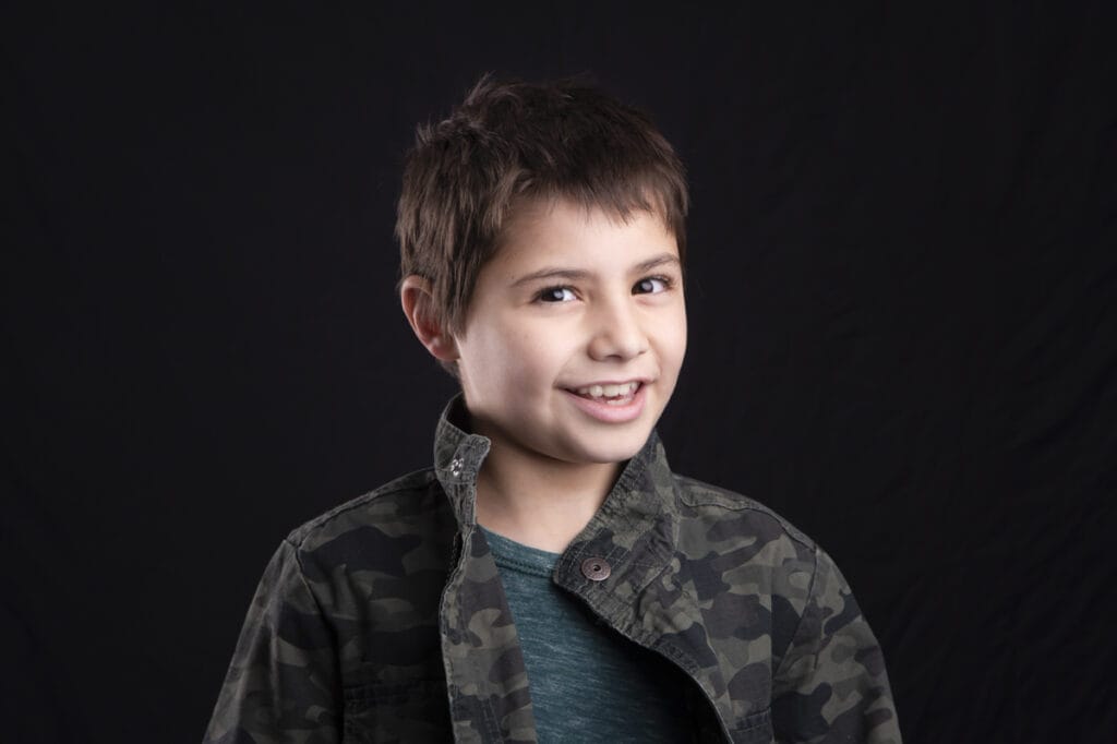 A young boy with short brown hair smiles while wearing a dark camouflage jacket over a teal shirt, posed against a plain black background—perfect for kids’ headshots in Calgary. - Jodi O Photography
