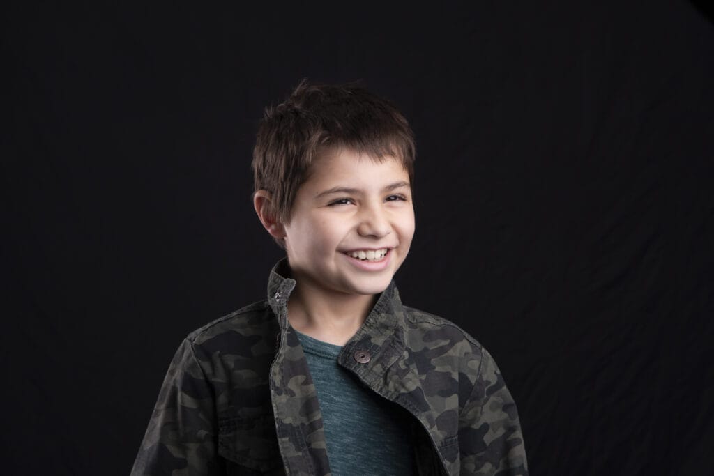 A young boy with short brown hair, wearing a camouflage jacket over a teal shirt, smiles and looks off camera against a plain black background—perfect for amazing kids headshots or Calgary kids photography. - Jodi O Photography