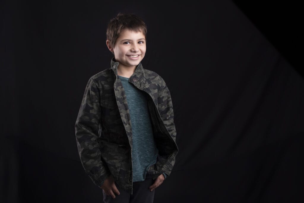 A young boy with short brown hair smiles, wearing a green camouflage jacket over a teal shirt, standing against a plain black background—perfect for kids’ headshots in Calgary. - Jodi O Photography