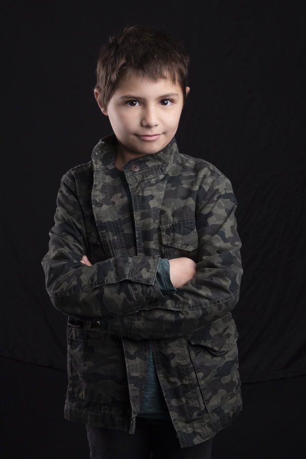 A young boy with short brown hair stands confidently with arms crossed, wearing a green camouflage jacket over a teal shirt, perfect for Calgary kids’ headshots, against a plain black background. - Jodi O Photography