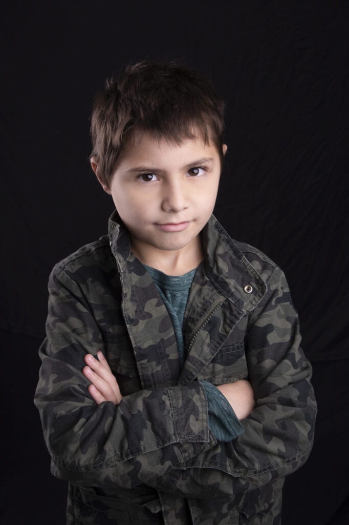 A young boy with short brown hair stands against a black background, wearing a green camo jacket over a green shirt. He looks at the camera with a slight smile and his arms crossed, perfect for Calgary kids headshots. - Jodi O Photography