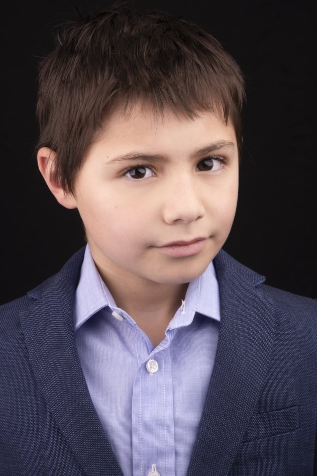 A young boy with short brown hair wears a blue blazer over a light blue shirt and looks confidently at the camera against a plain black background, perfect for kids’ headshots in Calgary. - Jodi O Photography