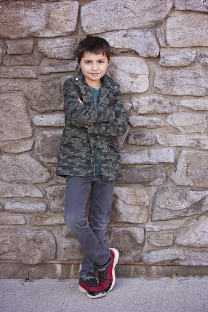 A young boy with short brown hair stands in front of a stone wall, arms crossed, wearing a camouflage jacket, teal shirt, gray pants, and red sneakers. He looks at the camera with a slight smile—perfect for kids headshots Calgary style. - Jodi O Photography