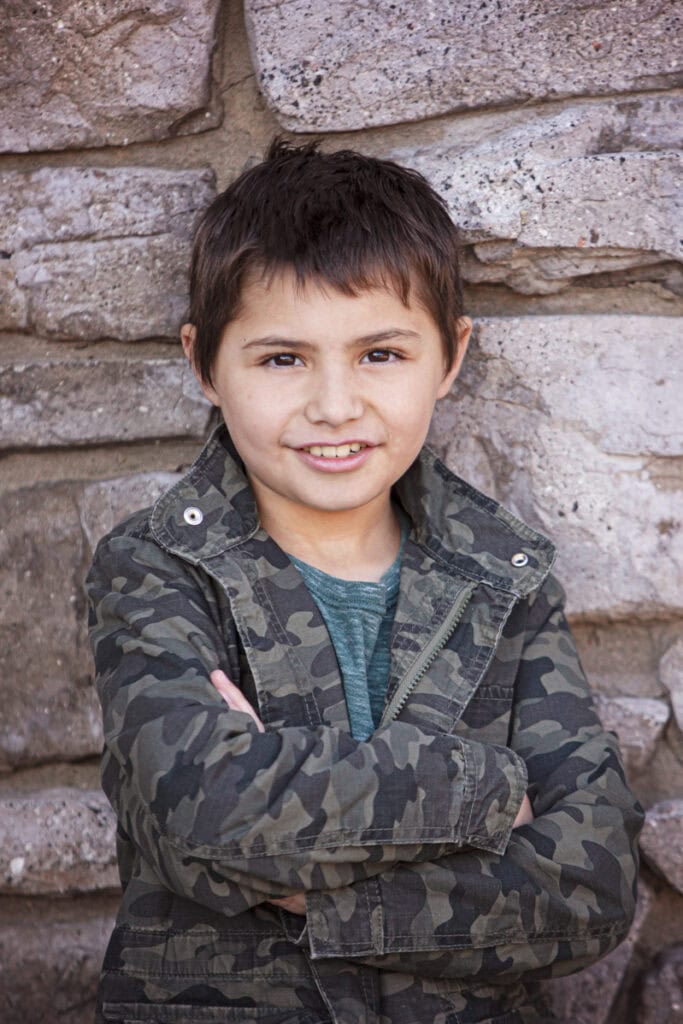 A young boy with short dark hair stands in front of a stone wall in Calgary, smiling with his arms crossed. He is wearing a green camo jacket over a green shirt, perfect for vibrant kids headshots. - Jodi O Photography