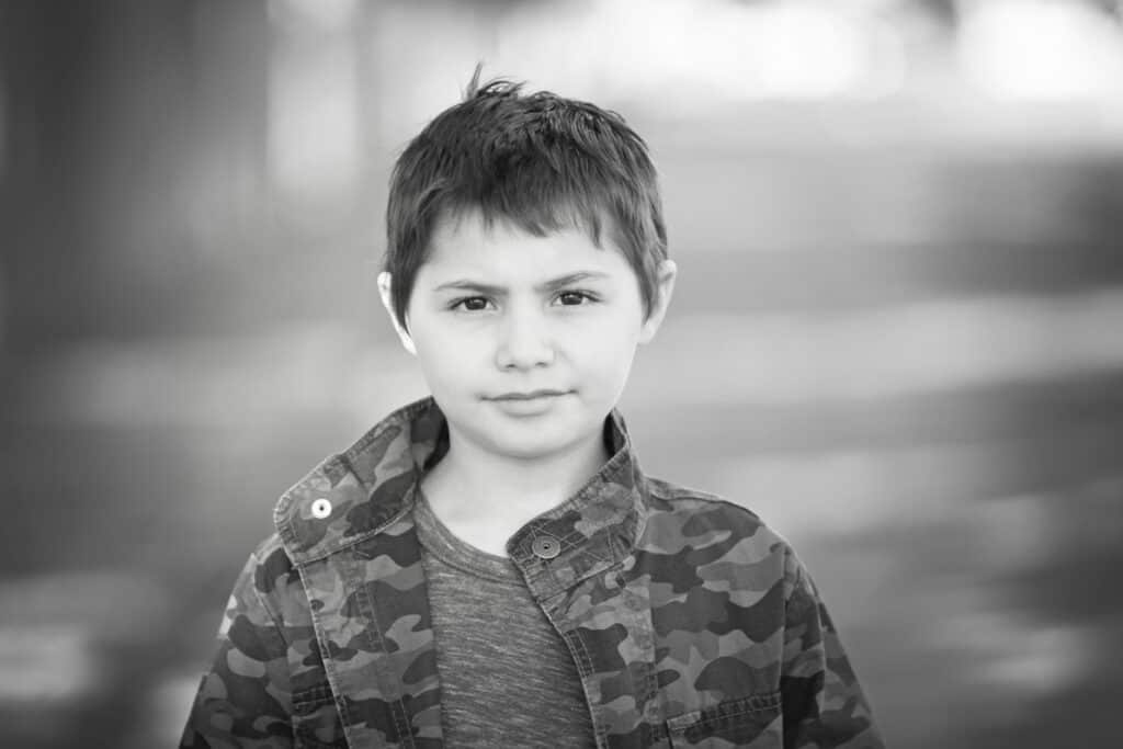 Black and white photo of a young boy with short hair, wearing a camouflage jacket and t-shirt, standing outdoors in Calgary. A striking example of kids headshots Calgary, with a neutral expression and blurred background. - Jodi O Photography
