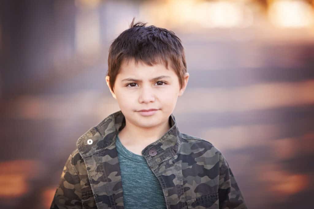 A young boy with short brown hair stands outdoors in Calgary, wearing a green shirt and camouflage jacket, looking directly at the camera with a neutral expression—perfect for kids headshots. The background is blurred with warm, soft lighting. - Jodi O Photography