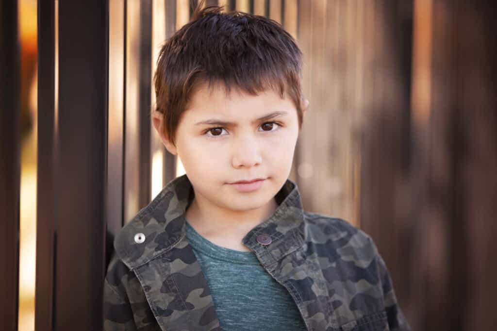 A young boy with short dark hair stands by a metal fence in Calgary, wearing a green camouflage jacket over a teal shirt. Looking calmly at the camera with a neutral expression, he embodies the essence of authentic kids headshots against a softly blurred background. - Jodi O Photography